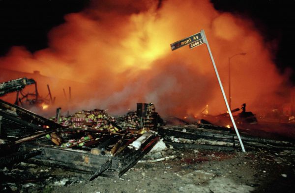 The remains of a commercial building smolder, as another building burns out of control, in Los Angeles, early on the morning of April 30, 1992, after riots broke out in response to the verdict in the Rodney King beating trial. Photo by Douglas C. Pizac/Associated Press.