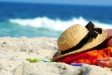 A person lies face down on a red towel at a beach, face hidden, with a hat covering the back of their head.
