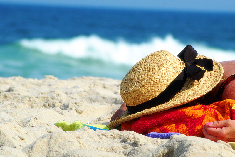 A person lies face down on a red towel at a beach, face hidden, with a hat covering the back of their head.