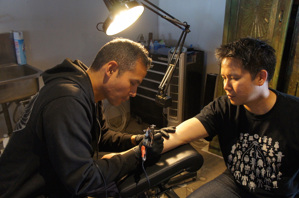 Ky-Phong Tran getting a tattoo with his right arm out while a tattoo artist leans in.