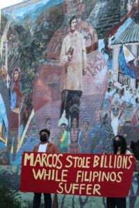 Two people wearing masks hold up a red banner with yellow words saying “Marcos Stole Billions While Filipinos Suffer” in front of the “Gintong Kasaysayan, Gintong Pamana (Filipino Americans: A Glorious History, A Golden Legacy)” mural at Unidad Park in Los Angeles.