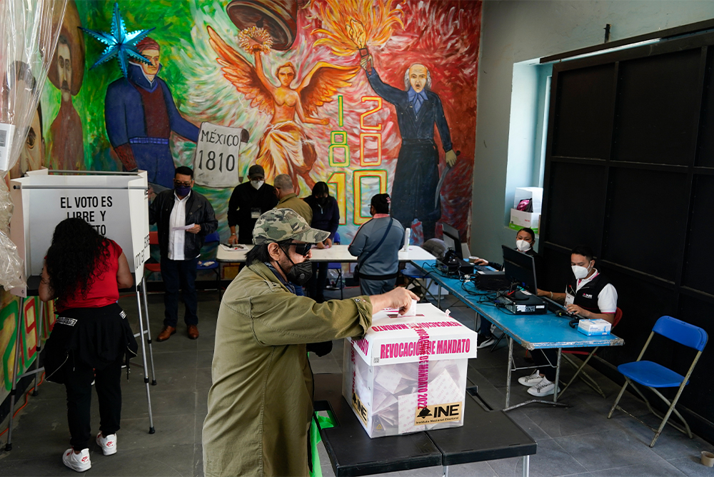A man in a mask and sunglasses cast a vote into a box. In the background, poll workers wearing masks sit at a blue table.