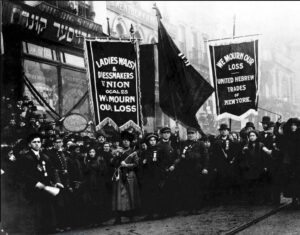 Black and white photo of a crowd of men and women holding large banners. One banner reads "Ladies waist & Dressmakers Union local 25 We mourn our loss" and another banner reads "We mourn our loss - United Hebrew Trades of New York."
