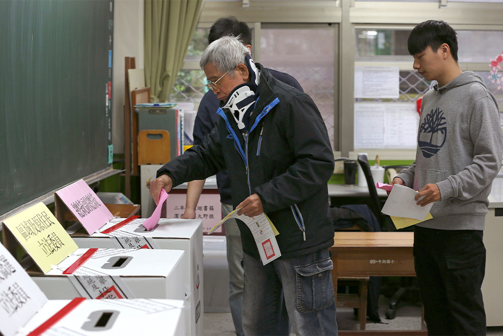 An elderly man with white hair and glass and wearing a black jacket places a pink sheet of paper into a white ballot box. His other hand holds two other pieces of paper, one white and and one yellow. A younger man in a gray sweater stands behind him holding his own papers.