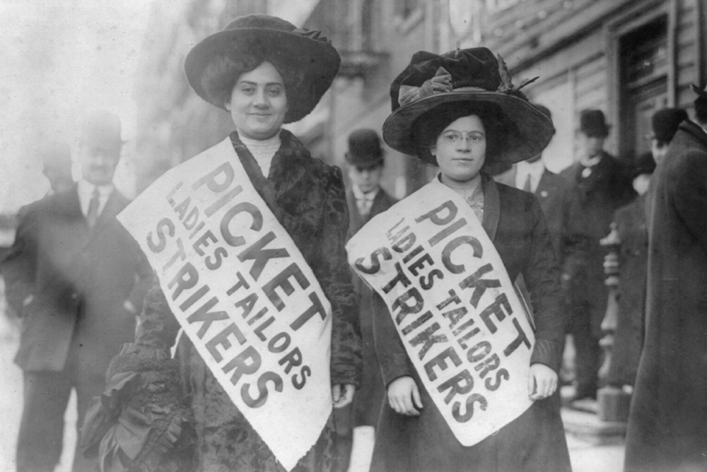 Black and white photo of two women standing side by side with banners over their bodies that reads "Picket Ladies Tailors Strikers"