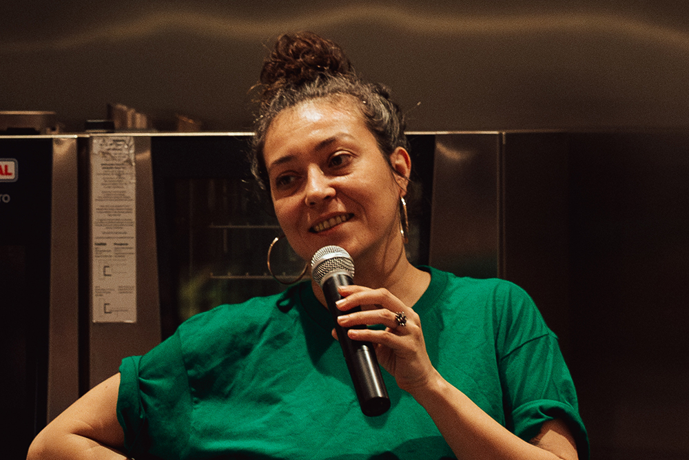 Zahara Gómez Lucini holds a microphone with mouth open in mid-speak. She wears a green t-shirt and stands in a kitchen, with an oven behind her.