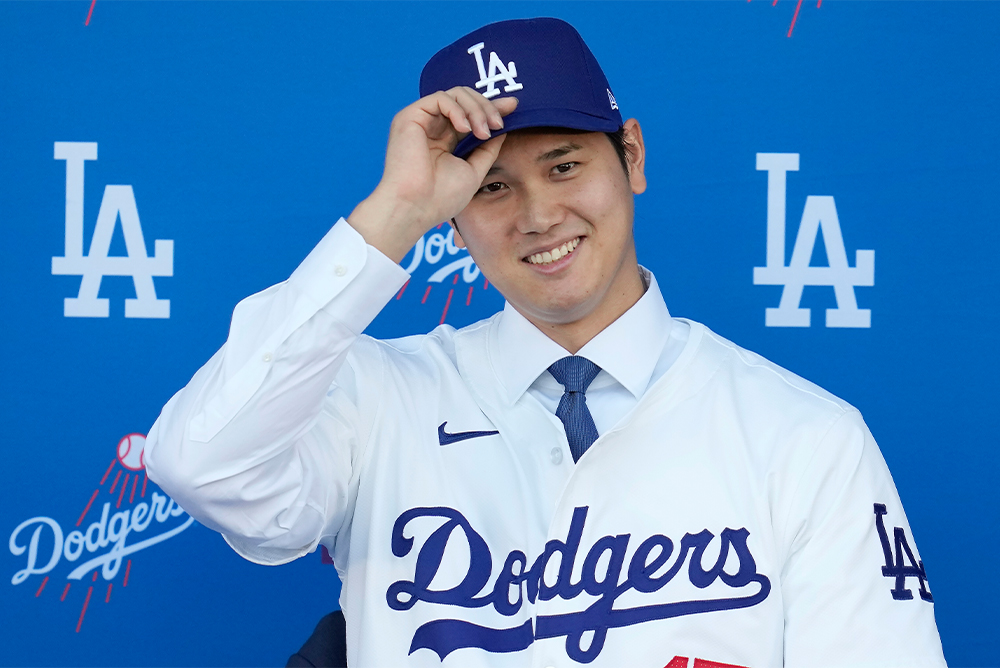 Baseball player Shohei Ohtani smiling at the camera. He wears a white Dodgers jersey and a blue L.A. cap.