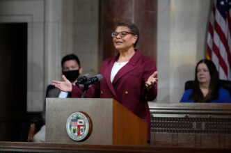 Karen Bass stands in front of a podium with both her hands raised in an open gesture at L.A. City Hall. Two council members are sitting and are visible in the background.