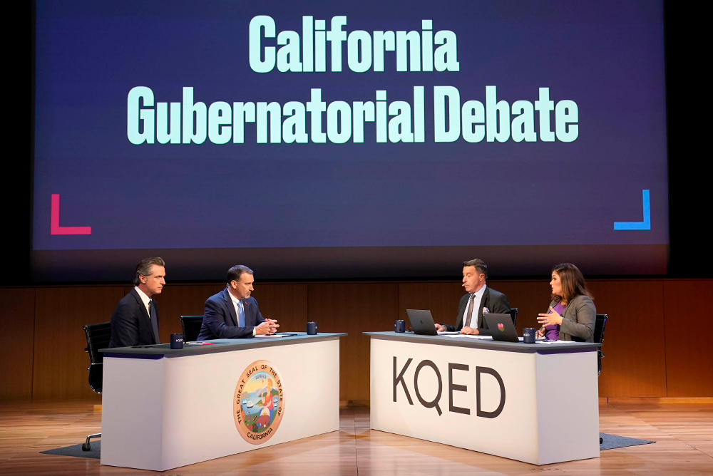 Two people, Governor Gavin Newsom and Republican challenger state senator Brian Dahle sit at the desk to the left. The desk has the California state symbol in front. On the desk on the right is Scott Shafer and Marisa Lagos. Their desk has the letters "KQED" on the front. The background large digital screen reads "California Gubernatorial Debate."